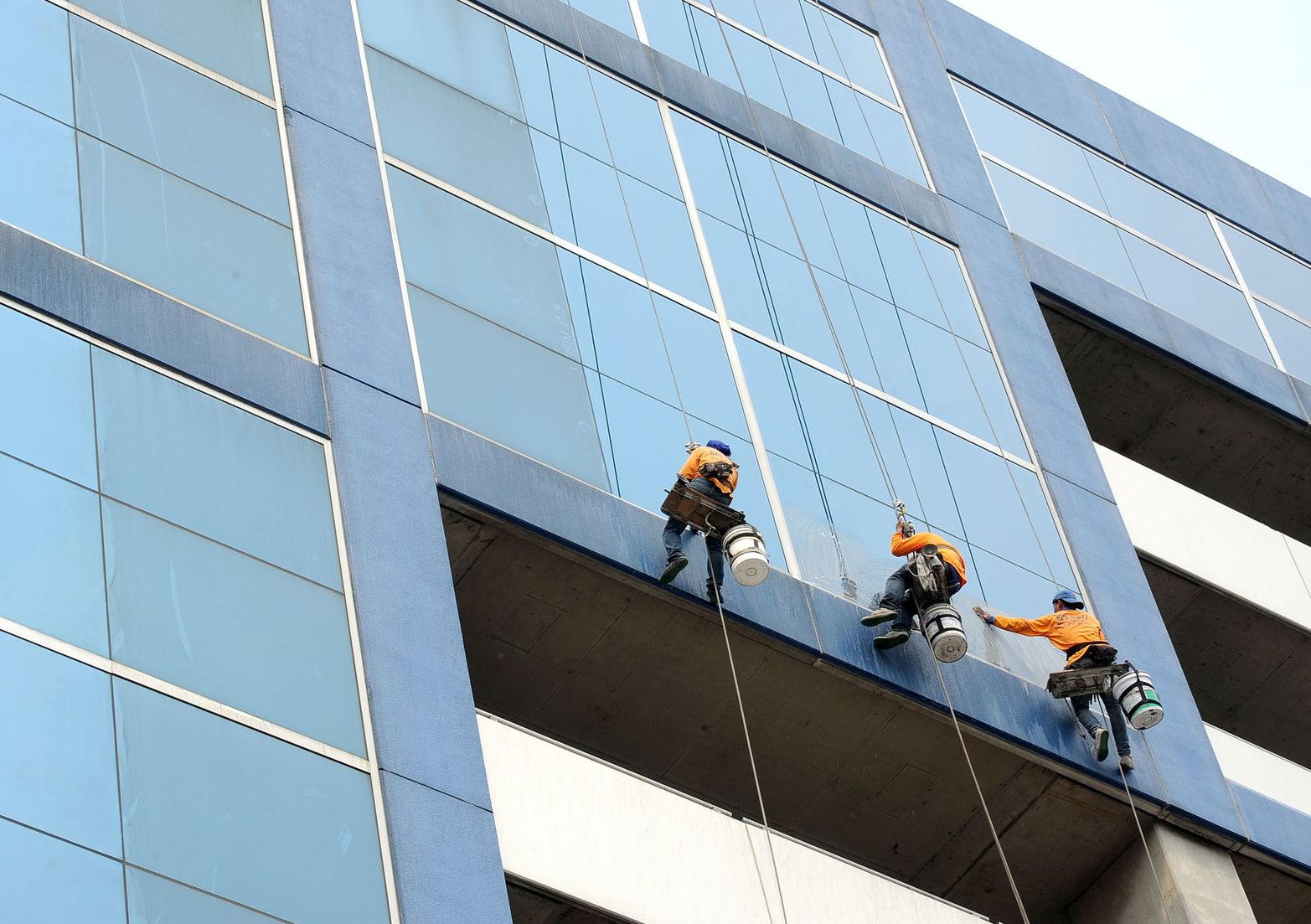 Drei Fensterputzer in orangefarbener Schutzkleidung reinigen die Glasfassade eines Hochhauses und hängen an Seilen